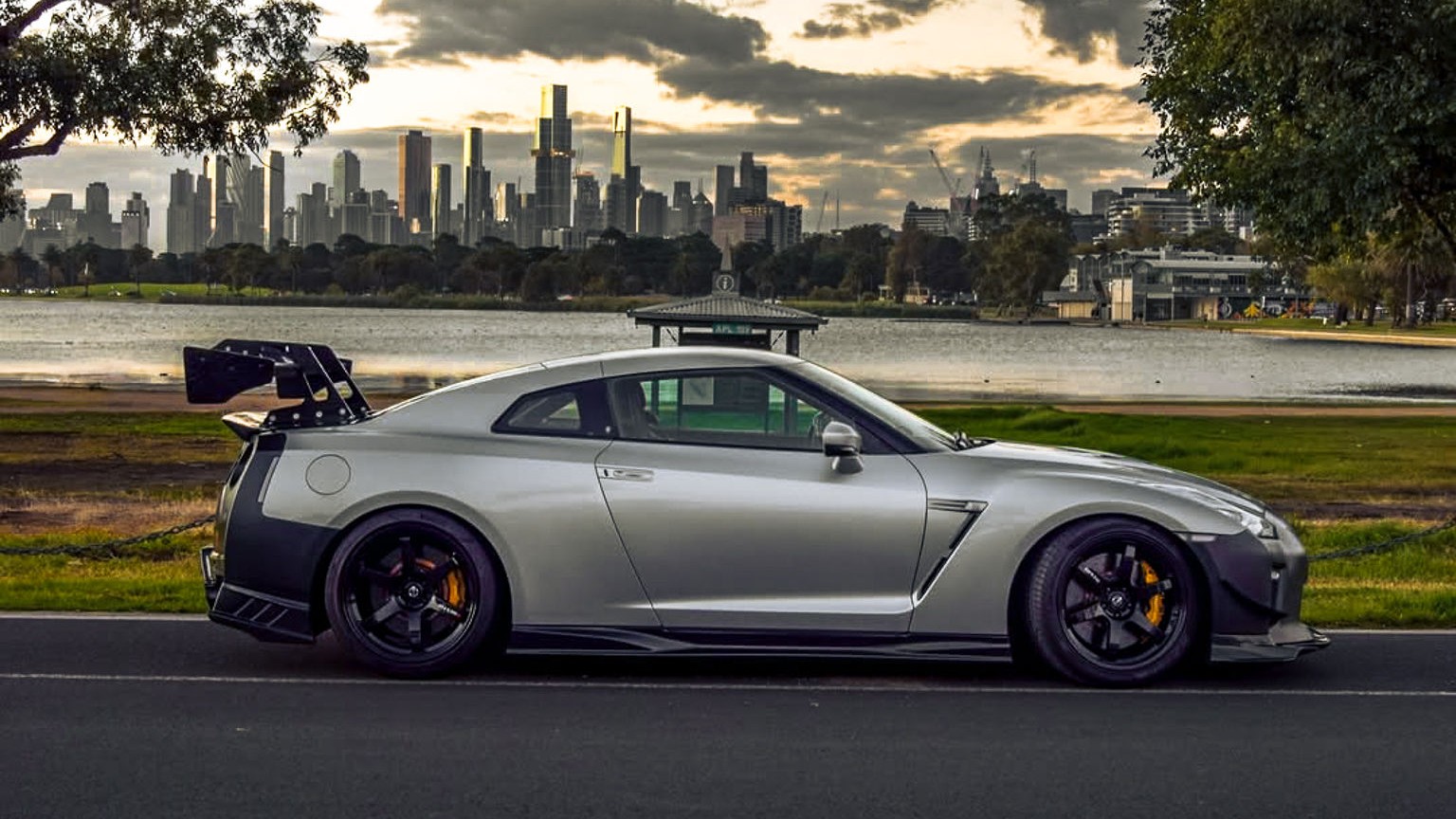 Silver Nissan GT-R parked by Albert Park lake with the Melbourne skyline at sunset